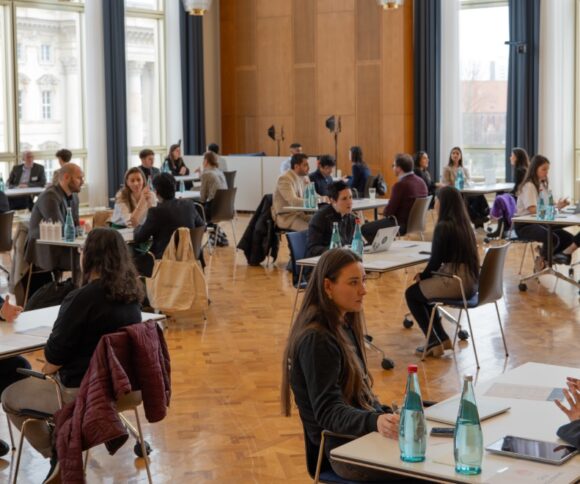 Das Bild zeigt einen großen Raum mit vielen Tischen, an denen jeweils zwei Personen im Gespräch sind.