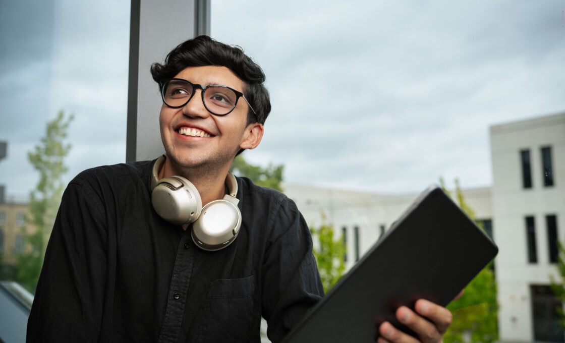 Male student with glasses and headphones smiling with a tablet in front of a modern building.