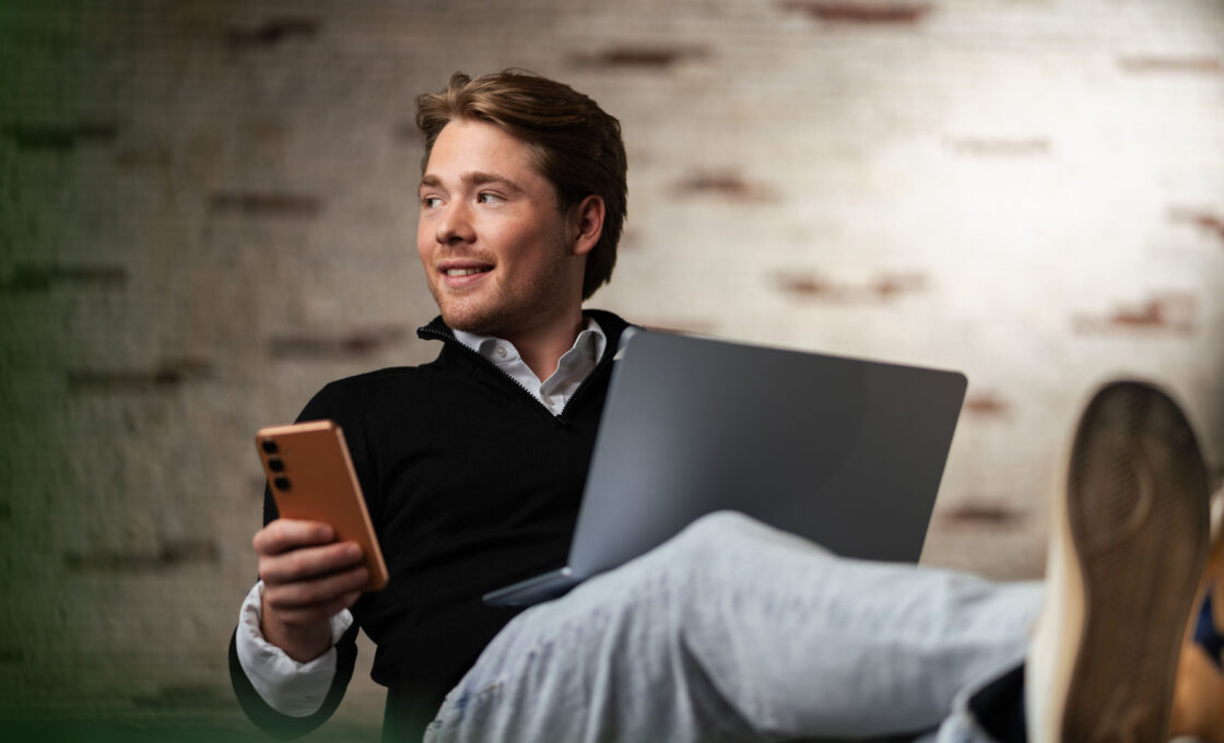 Student mit Smartphone in der Hand lehnt entspannt mit Laptop auf einem Sofa.