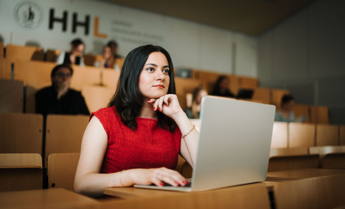 Female student in a red top sitting with a laptop in the lecture hall, pondering.