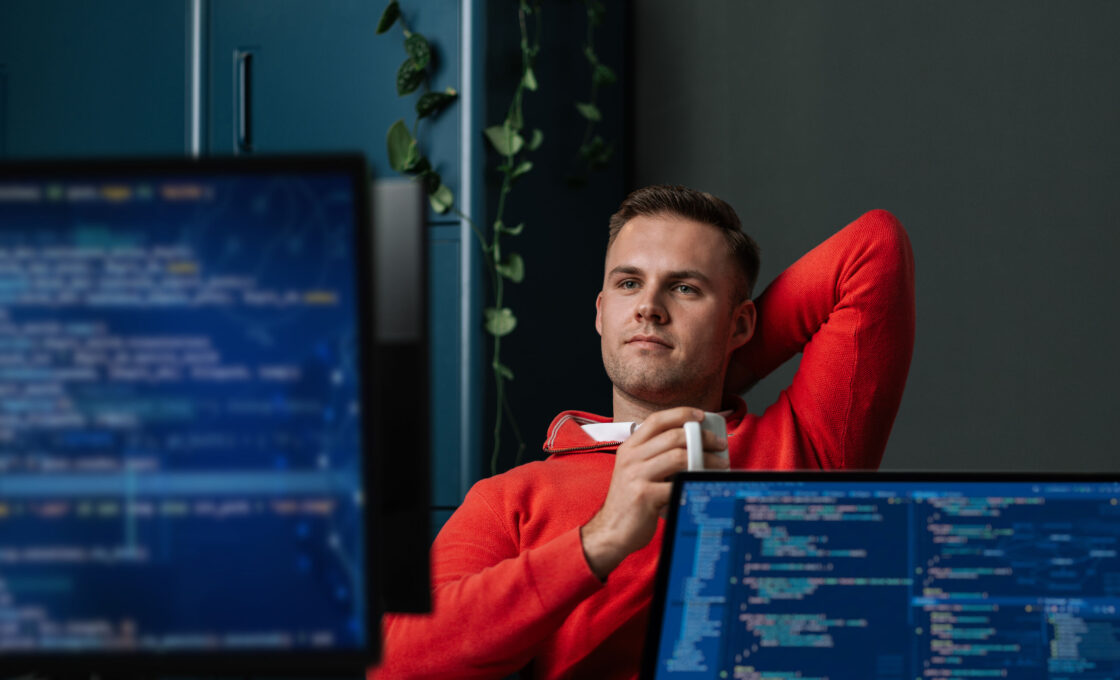 Male student in a red sweater sitting in front of screens showing code lines.