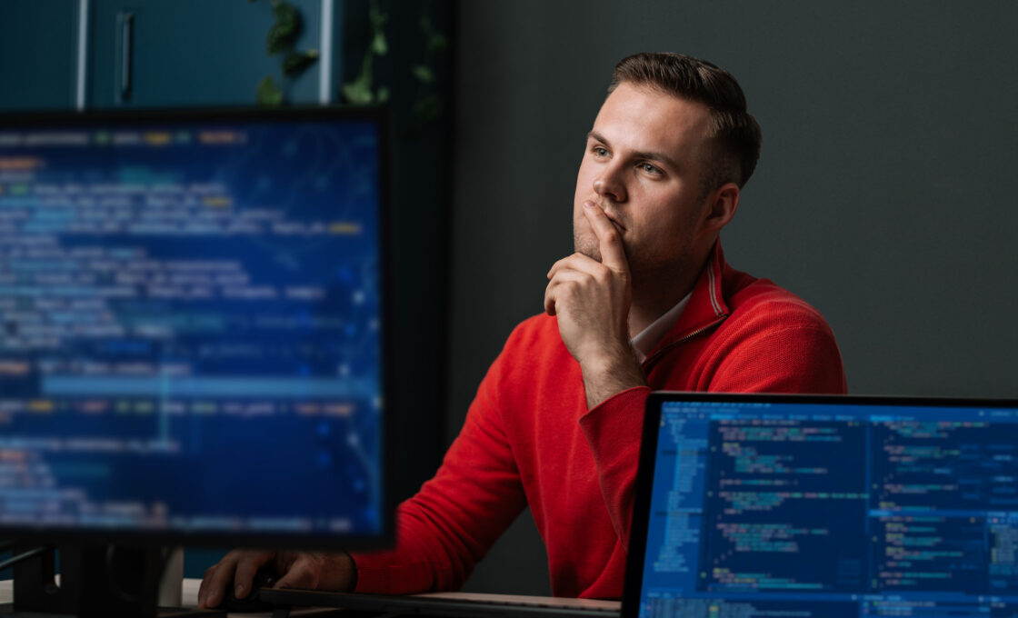 Student im roten Hoodie sitzt nachdenklich vor Monitor mit Programmcode.