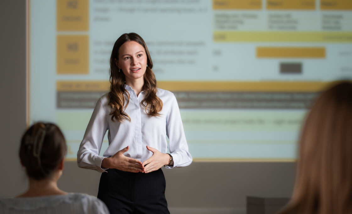 Studentin in weißer Bluse steht vor einer Präsentationsleinwand im Vorlesungssaal.