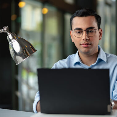 Male student with glasses working intently on a laptop in an office.