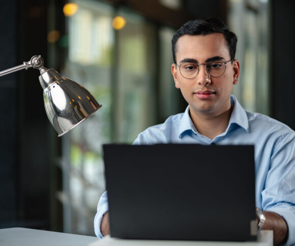 Male student with glasses working intently on a laptop in an office.
