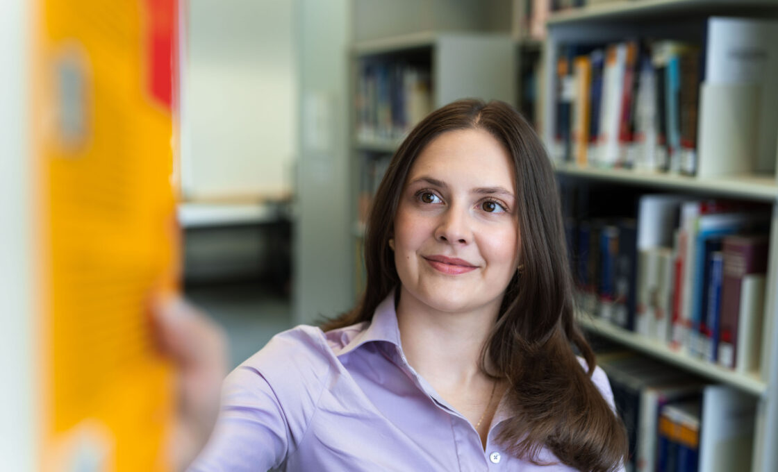 Female student reaching for a book on a library shelf and looking to the side.