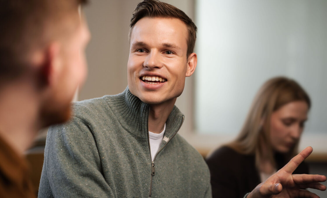 Male student in a grey sweater looking attentively toward a conversation partner.